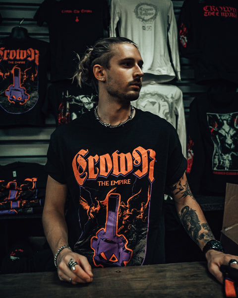 A person with tattoos and jewelry stands behind a counter, wearing a Crown the Empire shirt. Band merchandise featuring bold graphics and logos hangs on the wall behind them.