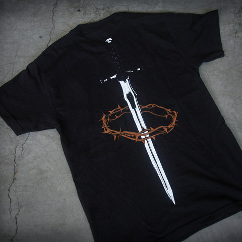 A black heavy cotton unisex Crown The Empire tee lies flat on concrete, featuring a large downward-pointing sword graphic with a brown crown of thorns encircling the blade near the hilt.