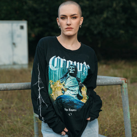 A person with a shaved head and hoop earrings leans on a metal railing outdoors, wearing a Crown The Empire unisex long sleeve black graphic shirt with a grim reaper design, made from 100% combed cotton; trees and a white utility box are behind.