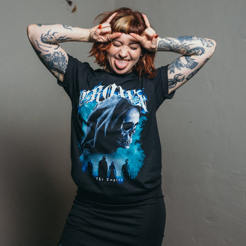 A tattooed woman playfully sticks out her tongue and makes a peace sign, wearing a Crown The Empire AS Colour Staple Tee (regular fit, 100% combed cotton) with a blue hooded skull design and a black skirt, set against a gray background.