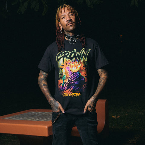 A person with red and blonde dreadlocks and facial piercings stands outdoors at night, wearing a black Crown The Empire tee featuring a colorful CROWN graphic. A picnic table is visible in the background.