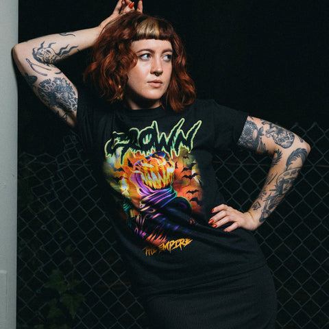 A red-haired, tattooed woman poses by a chain-link fence in a black Crown The Empire AS Colour Staple Tee with 100% combed cotton, featuring a colorful spooky graphic of "CROWN" and a sinister pumpkin-headed figure.