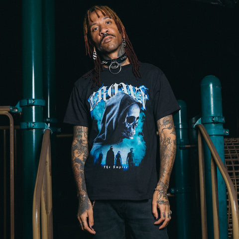 A person with reddish-brown dreadlocks and facial piercings stands on a playground at night, wearing a Crown The Empire tee with a bold skull graphic, black pants, and a thick choker necklace.