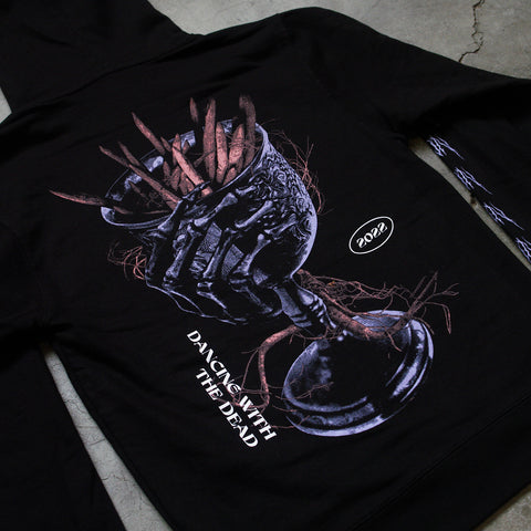 Up close Photo of the back of a black hoodie on a grey background. The sleeves say Crown on them repeatedly in a death metal font in purple. The back of the hoodie features a purple goblet with a skeleton hand wrapped around it. In white are the words dancing with the dead and 2022.