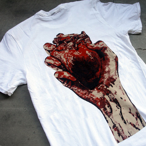close up, angled image of the back of a white tee shirt laid flat on a concrete floor. the tee has a full back print of a hand holding a bloody heart