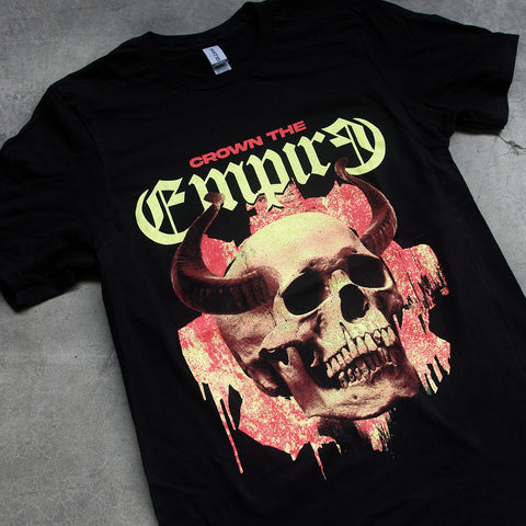 close up, angled image of a black tee shirt laid flat on a concrete floor. tee has full body print of a skull with horns. at the top says crown the empire