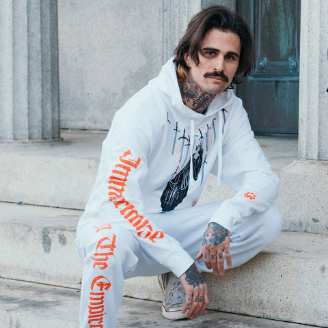 image of a white pullover hoodie worn by a man kneeling on steps outside. hoodie has full body print of a skeleton in a cloak with swords surrounding it. the hood has a red print that says crown. the left sleeve says immortalize and the right cuff has the crown emblem logo