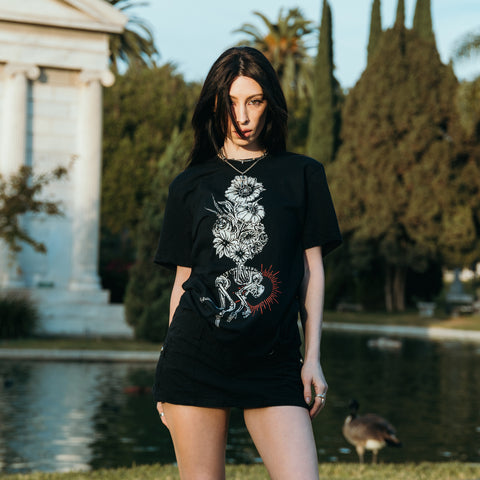 image of the front of a black tee shirt worn by a woman standing in front of a water. tee has a full body print of a bouquet of flowered above a hunched over skeleton.