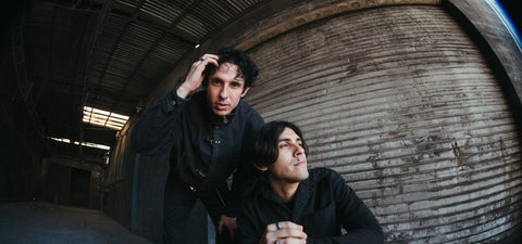 Two men dressed in black, one standing and touching his head, the other squatting with hands clasped, pose in front of a large, worn metal garage door in an industrial, dimly lit setting.