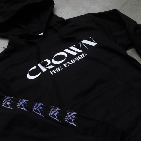  Up close Photo of the front of a black hoodie on a grey background. The front of the hoodie says crown the empire in white. The sleeves say Crown on them repeatedly in a death metal font in purple