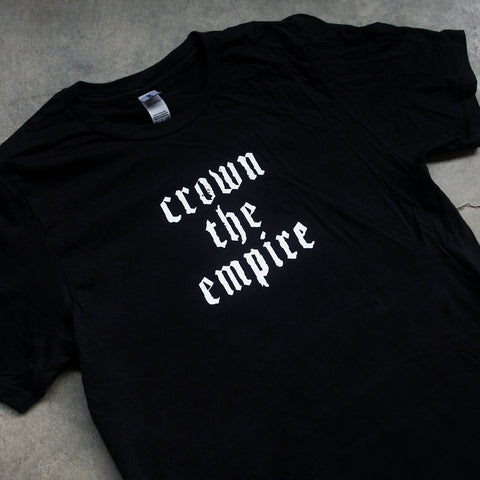 close up, angled image of the front of a black tee shirt laid flat on a concrete floor. the tee has a small center chest print in white that says crown the empire in stacked text. 