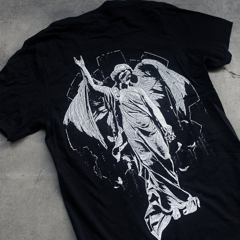 close up, angled image of the back of a black tee shirt laid flat on a concrete floor. tee has a full back print of an angel with one arm raised.