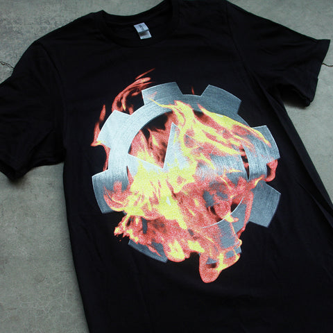 close up, angled image of the front of a black tee shirt laid flat on a concrete floor. tee has a full chest print of a cog on fire. 
