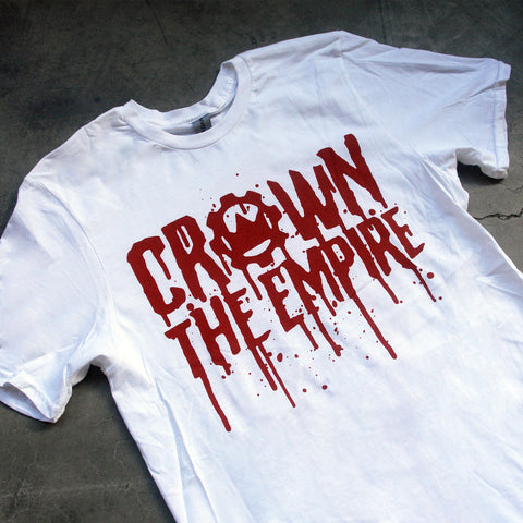 close up, angled image of the front of a white tee shirt laid flat on a concrete floor. the tee has a full print in red across the chest that says crown the empire in blood dripping text.