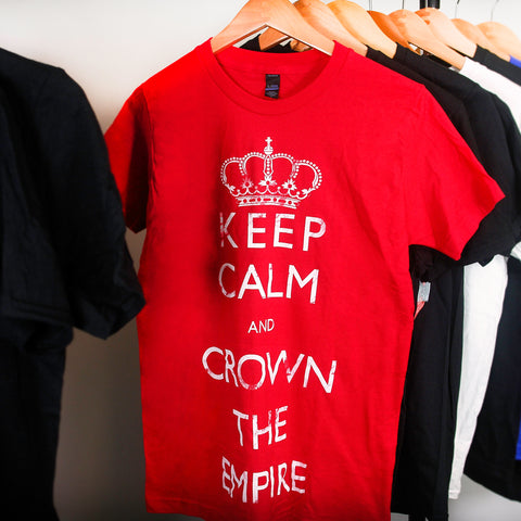 Image of a red tshirt on a hanging clothes rack in front of a white background. The tshirt has an image of a crown at the top and below that in white writing the text says keep calm and crown the empire.