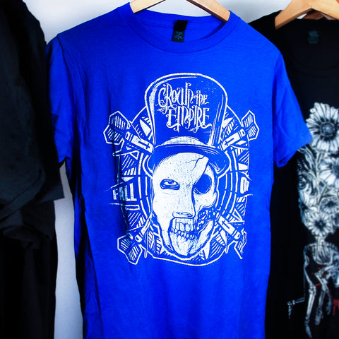 Royal blue tee on a hanger in front of a  white background. The tshirt features a mans face that is half skeletal. He is wearing a top hat that says crown the empire. His face is on a dart board and surrounded by darts. Next to his face are the words fall out. This design is done in white color.