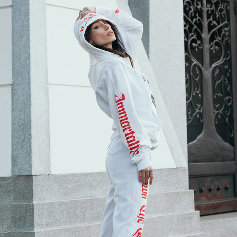 image of a white pullover hoodie worn by a woman standing outside. hoodie has full body print of a skeleton in a cloak with swords surrounding it. the hood has a red print that says crown. the left sleeve says immortalize and the right cuff has the crown emblem logo