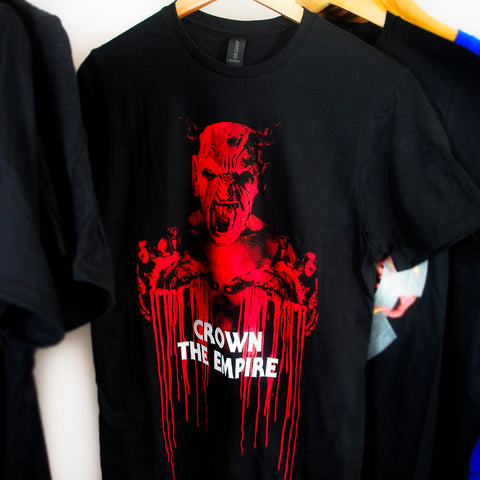 image of a black tee shirt on hanging in front of a white background. tee has full body print in red of a demon with bloody hands stretched out. over the image in white says crown the empire