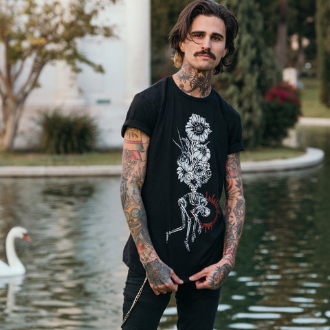image of the front of a black tee shirt worn by a man with tattoos standing in front of a water. tee has a full body print of a bouquet of flowered above a hunched over skeleton.